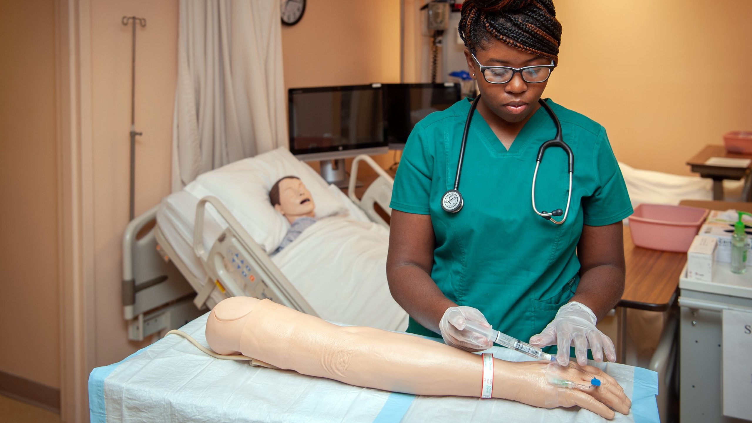 Female student in nursing simulation lab using syringe and tube on mannequin arm.