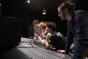 Four students standing and sitting in front of recording studio console, working on a project.