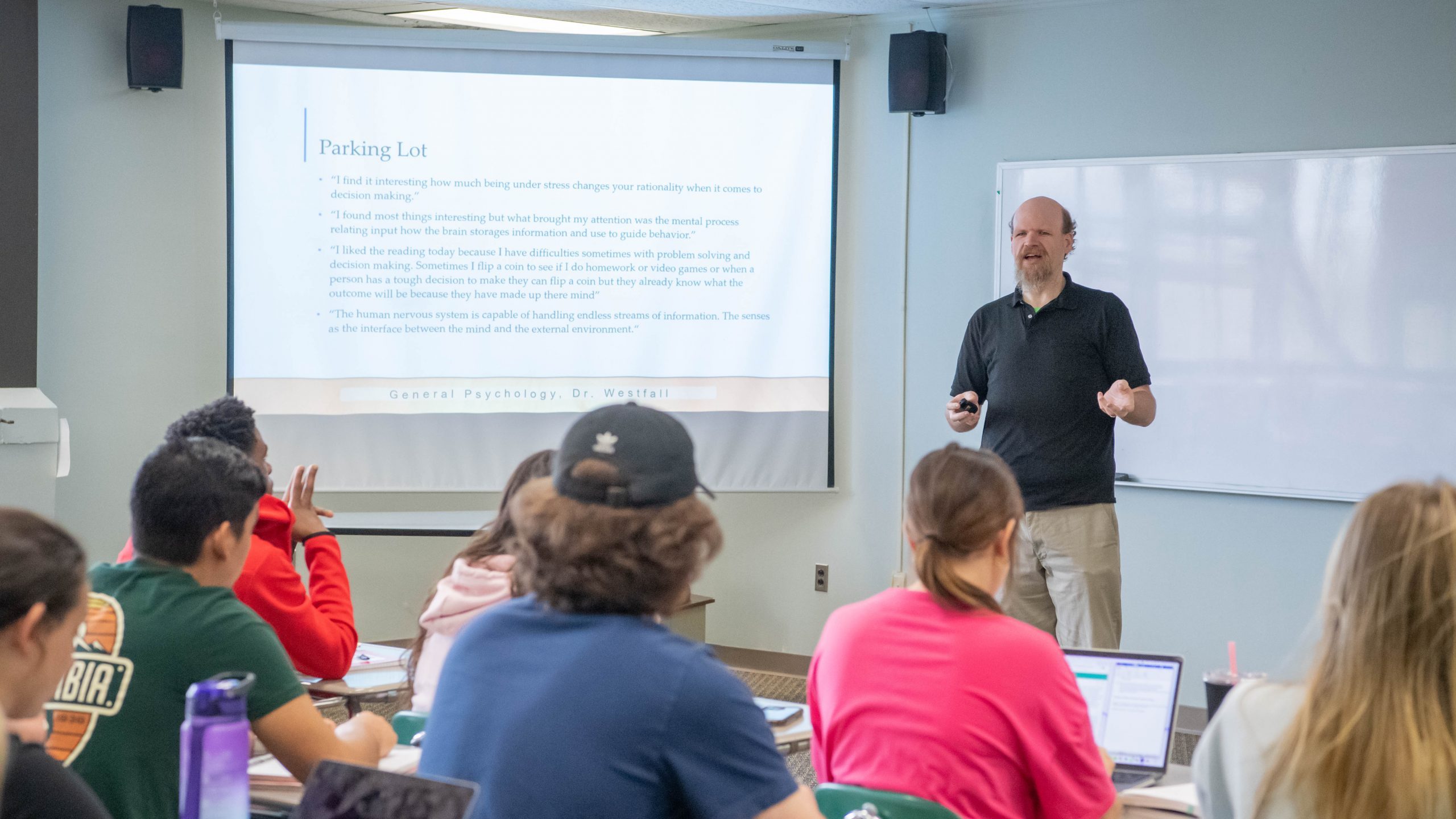 Psychology professor standing before class and teaching from a PowerPoint presentation that's displayed on a projector screen before the students.