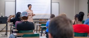 Professor teaching management class with PowerPoint presentation behind her.