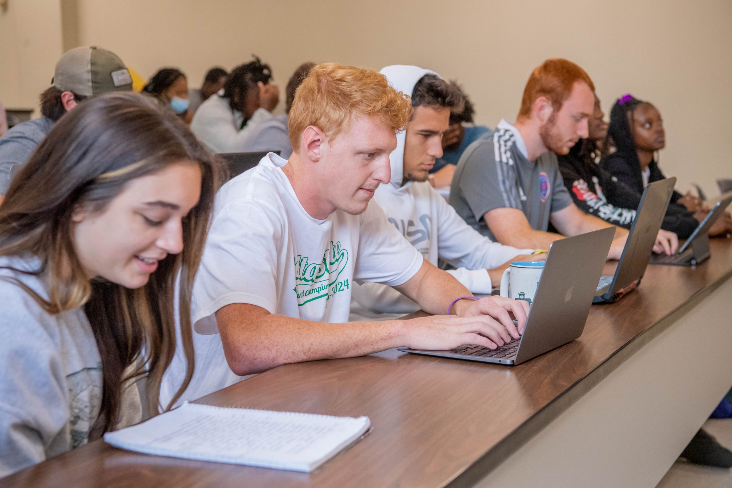 A front row of six students in a classroom taking notes on laptops and on paper.