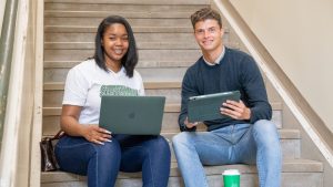Two students, one with laptop and one with an ipad, sitting on stairs smiling.