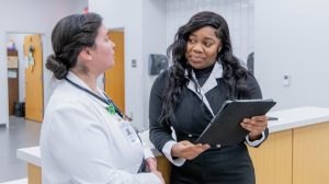 Two women talk in a professional setting; one in a lab coat and stethoscope, the other holding a tablet and dressed in business attire, discussing something work-related.