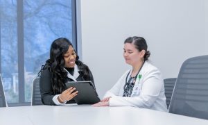 Two women sit at a conference table, one in business attire showing something on a tablet to the other in a lab coast, who listens attentively.