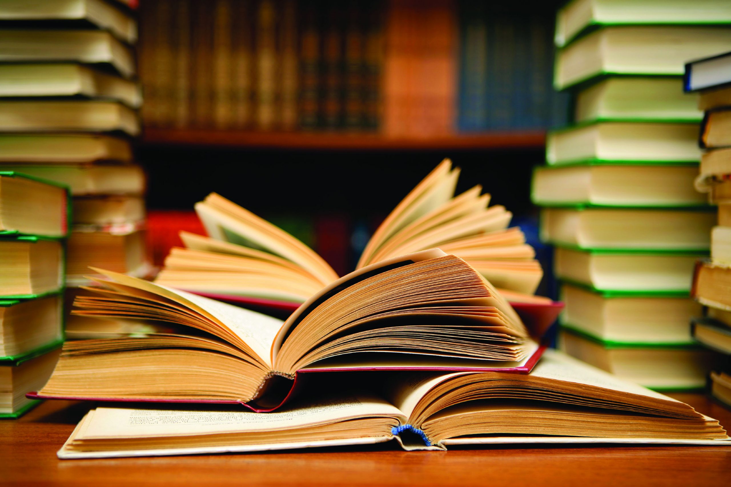 Books open on a table, surrounded by other books stacked on the same table and in a bookshelf behind the table.