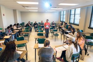 Students surrounding professor in a classroom setting while professor is teaching.