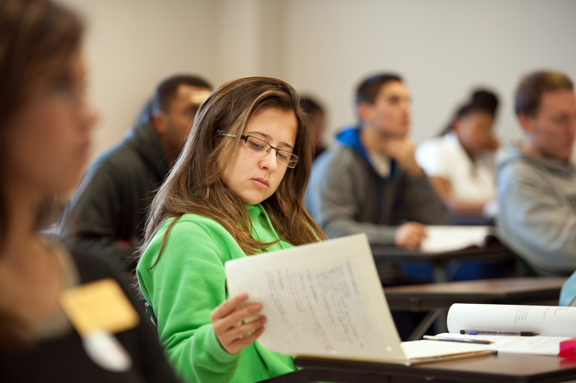 Student sitting at desk in a full classroom, reviewing written notes.