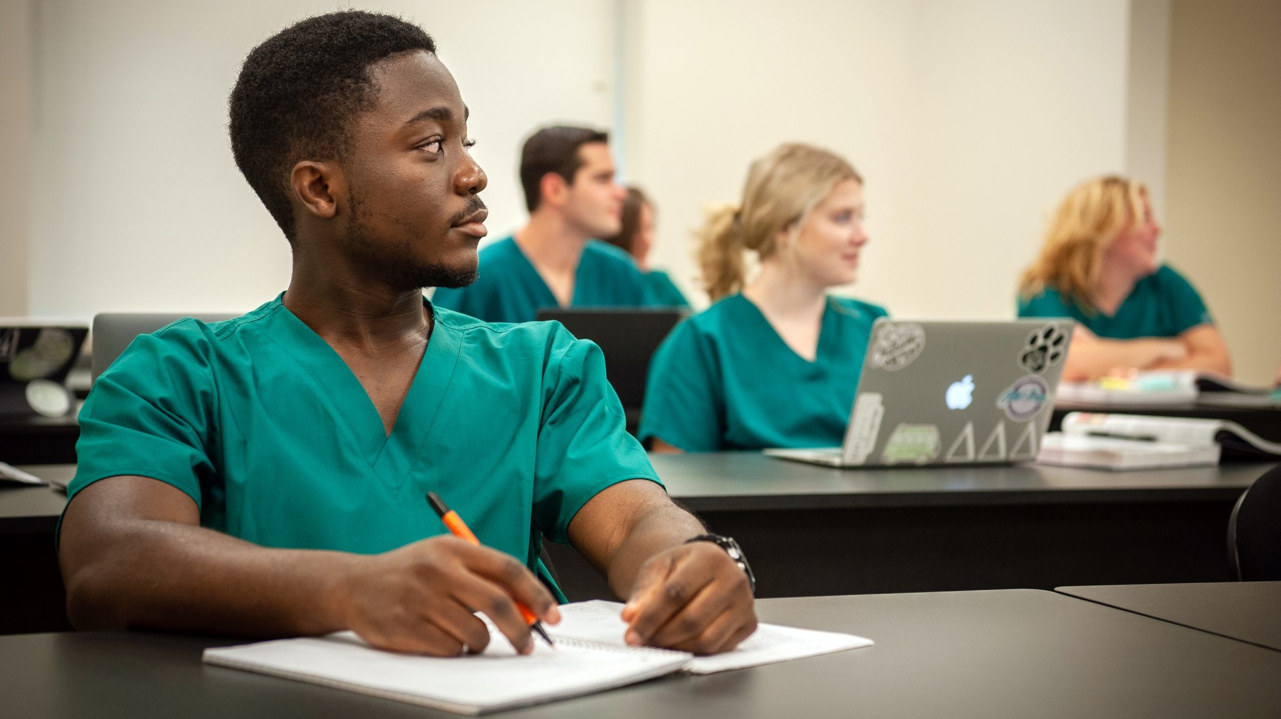 Student sitting at a table in class, taking notes during lecture, with four other students behind him.