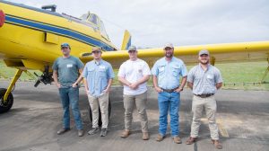 Five students standing in front of a crop duster plane on an airport runway.