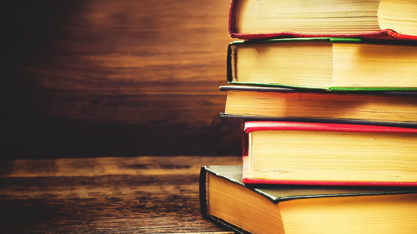 Five books stacked on a wooden table with a wooden wall in the background.