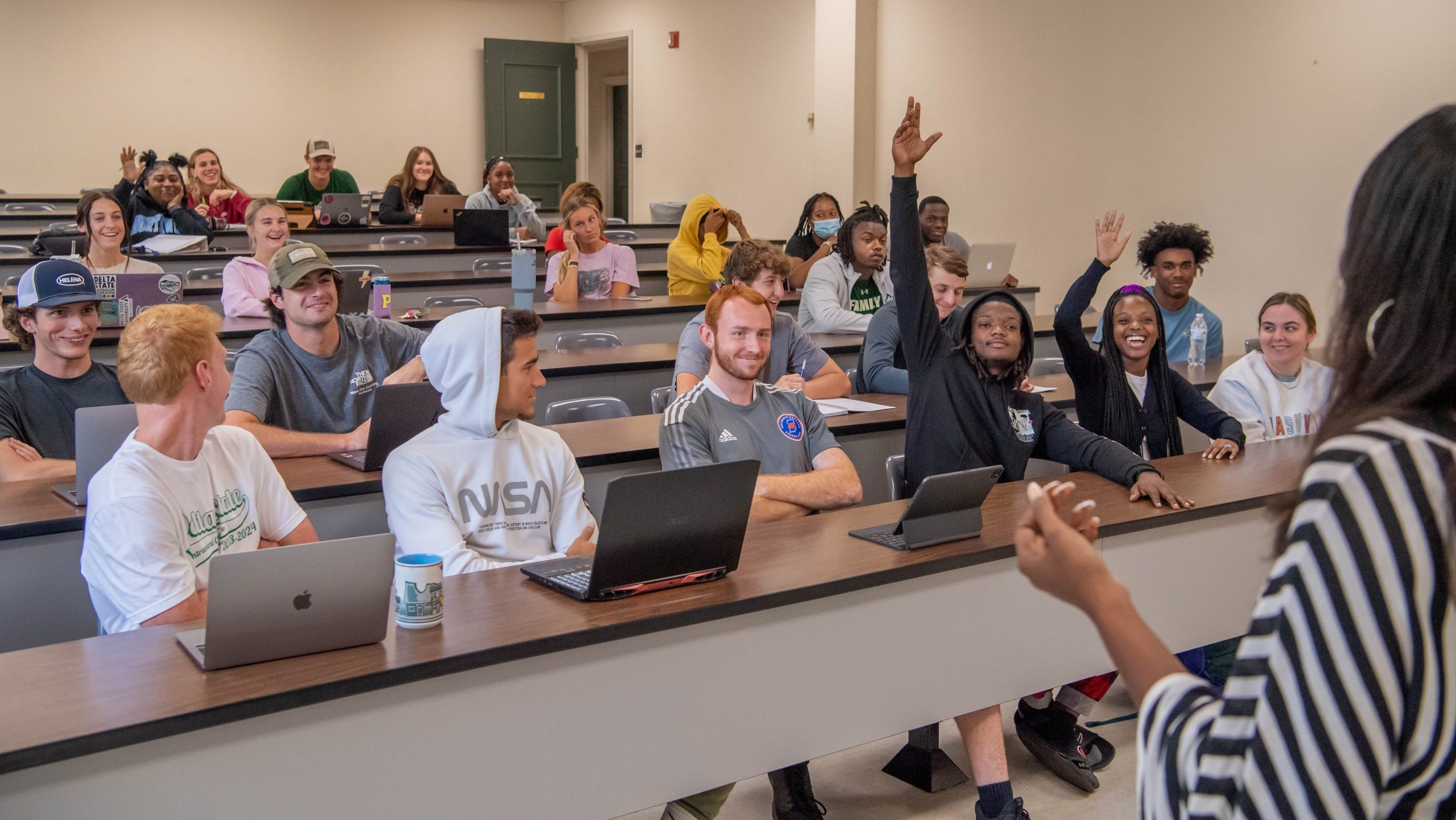 Two students on the front row of a full classroom, each with a hand raised to answer a question.