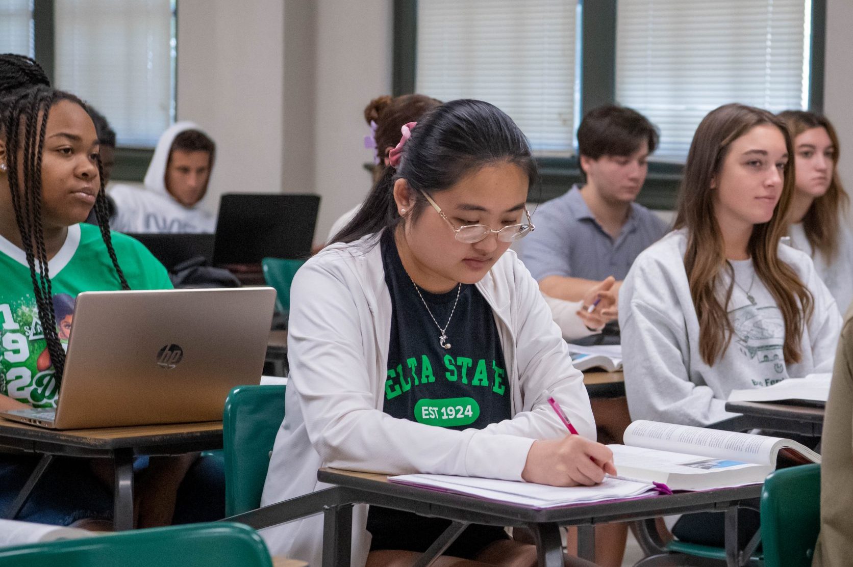 Student sitting at a desk in a full classroom, taking notes on paper with textbook open.