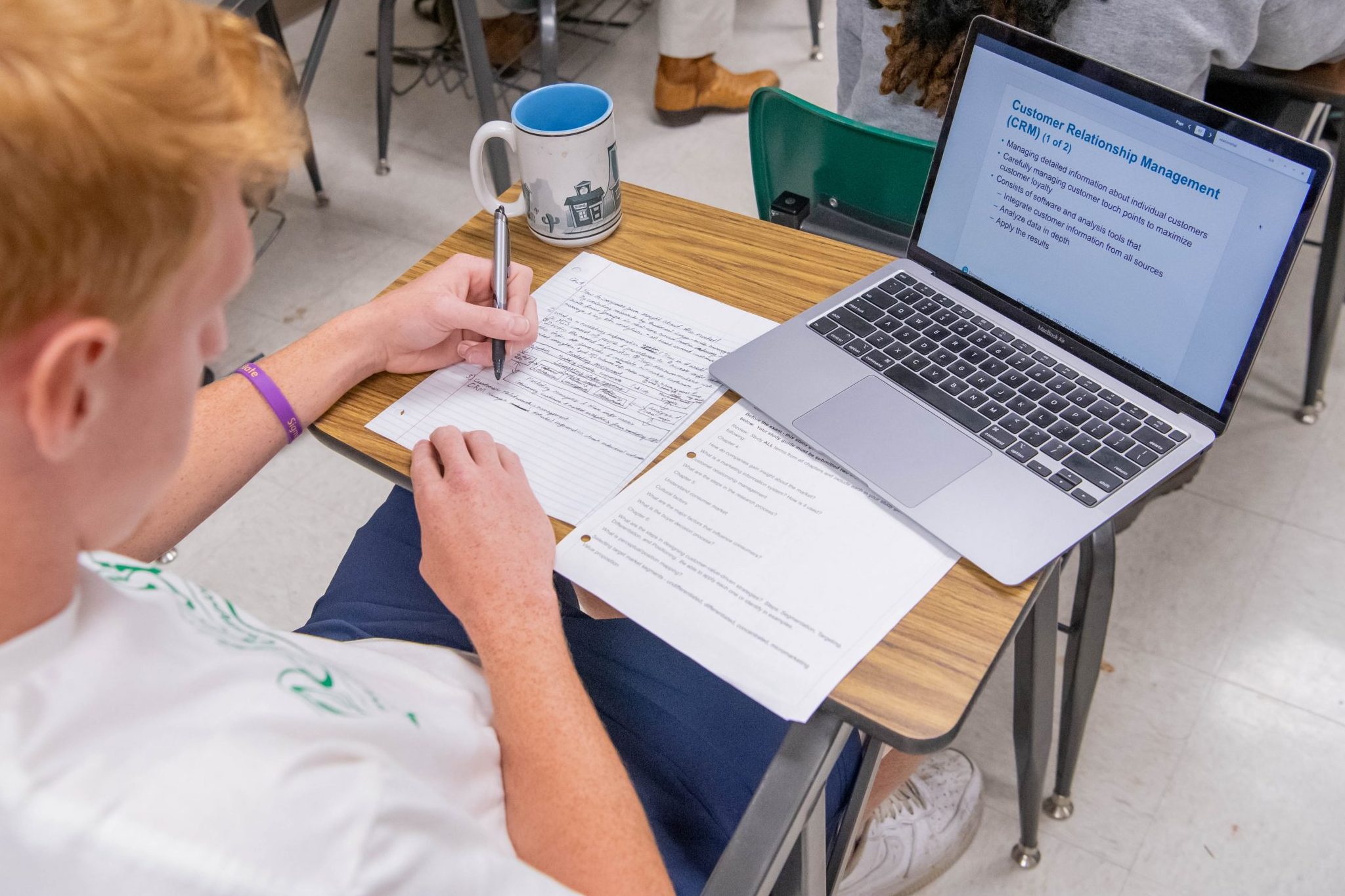 Student sitting at a desk in a classroom, taking notes on paper, and looking at a PowerPoint presentation open on his laptop.