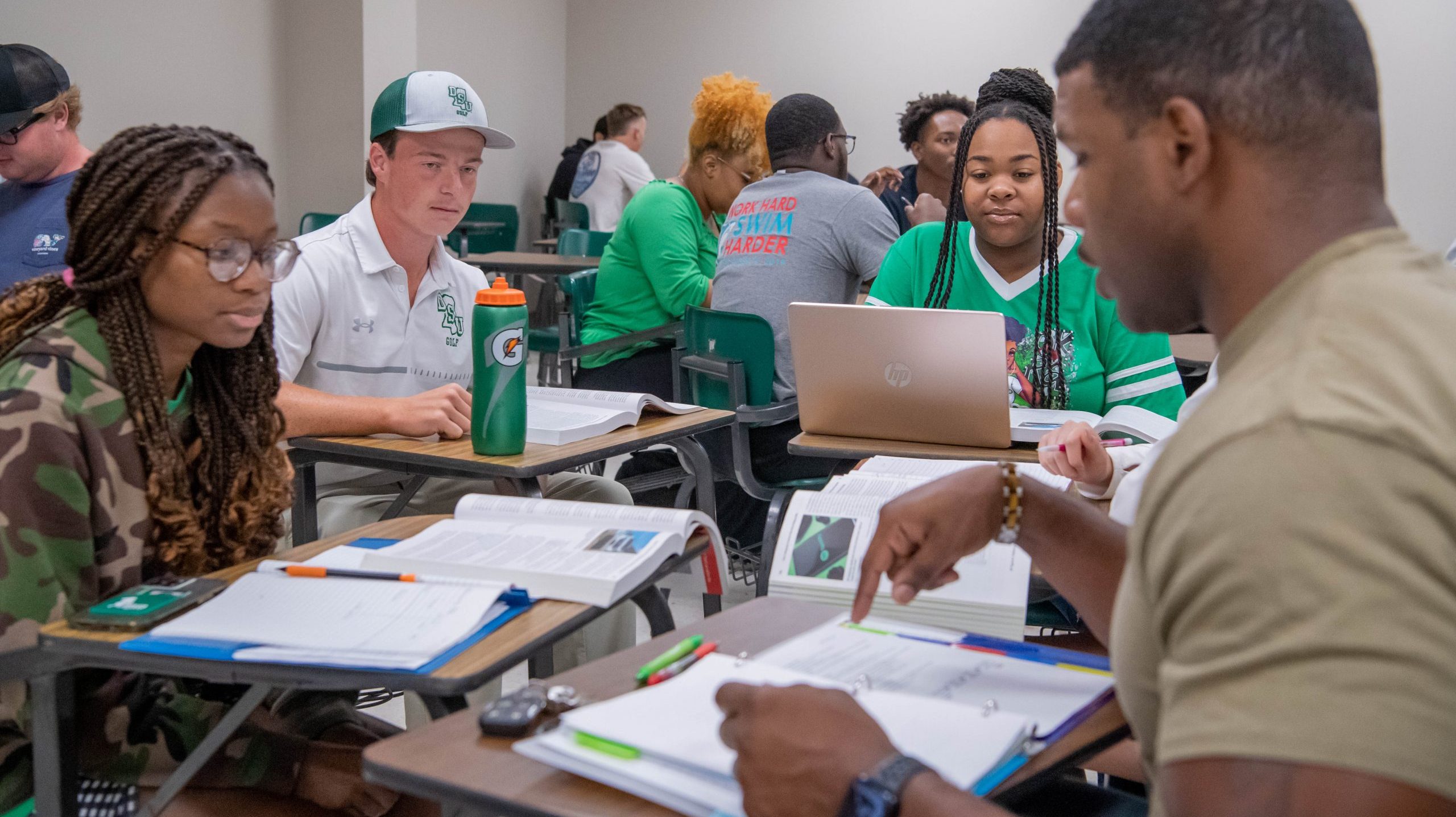 A group of four students working together on an assignment with other students in the background.