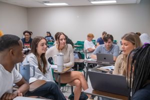 Group of six students working on assignment together during class, with other students in the background.
