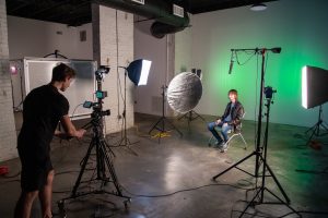 Student sitting in a chair with video equipment and lighting placed around him, while another student operates a camera on a tripod.