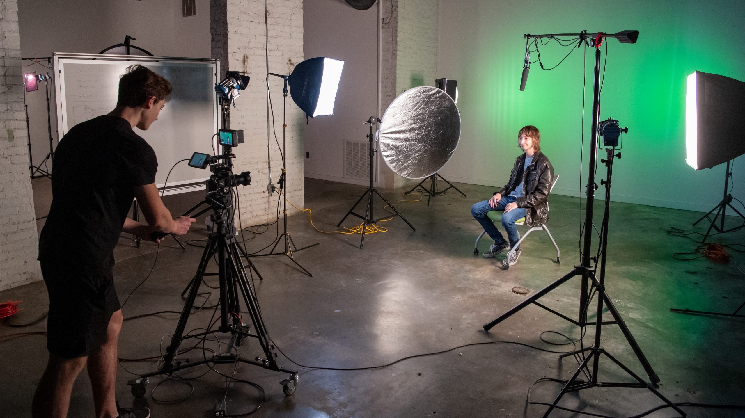 Student sitting in a chair with video equipment and lighting placed around him, while another student operates a camera on a tripod.