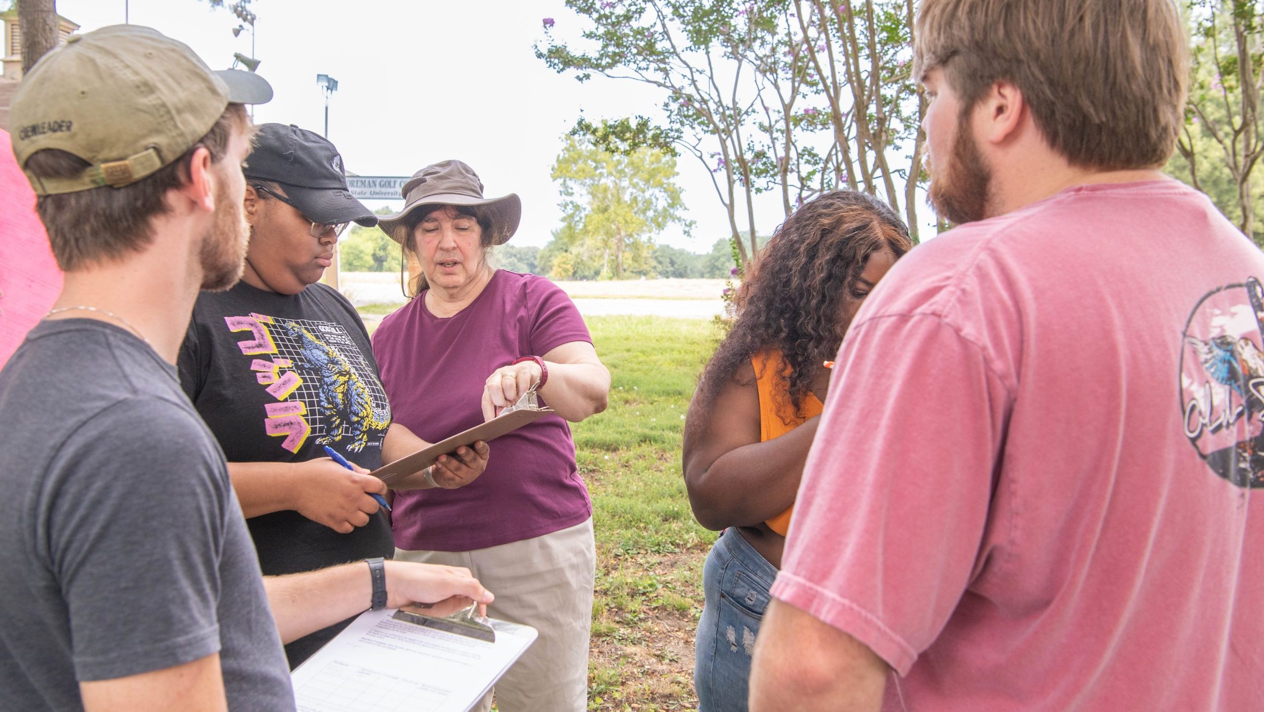 Professor with four students outside writing and reviewing data on clipboards.