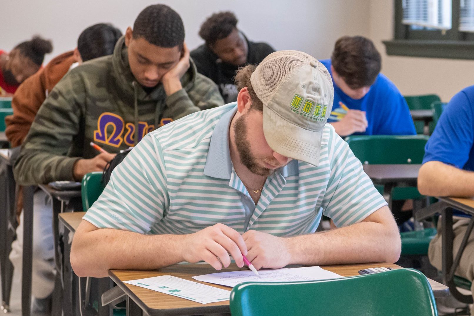 Student sitting at a desk completing assignment during class, with another six students behind him also at desks completing the assignment.