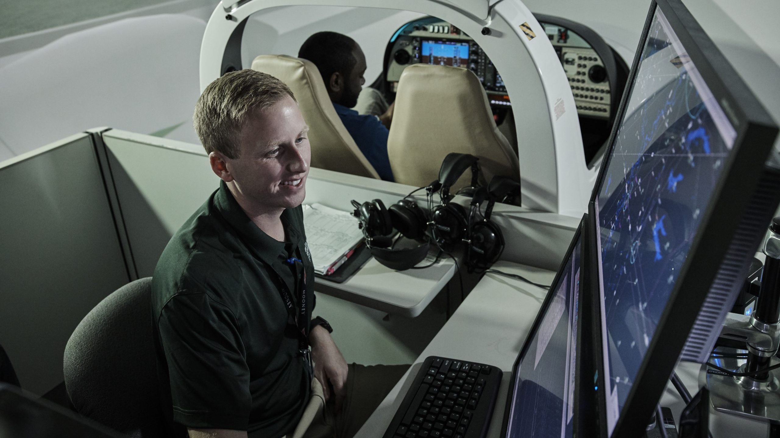 Student looking at computer screens, monitoring air traffic, while another student is in the flight simulator behind him.