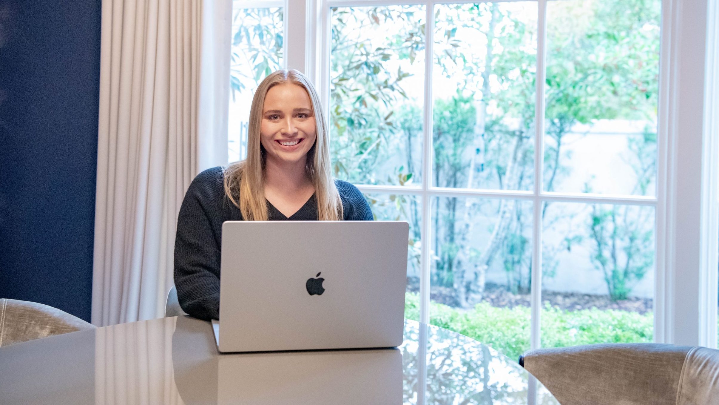 Student sitting at dining room table, with laptop and large window behind her at home.