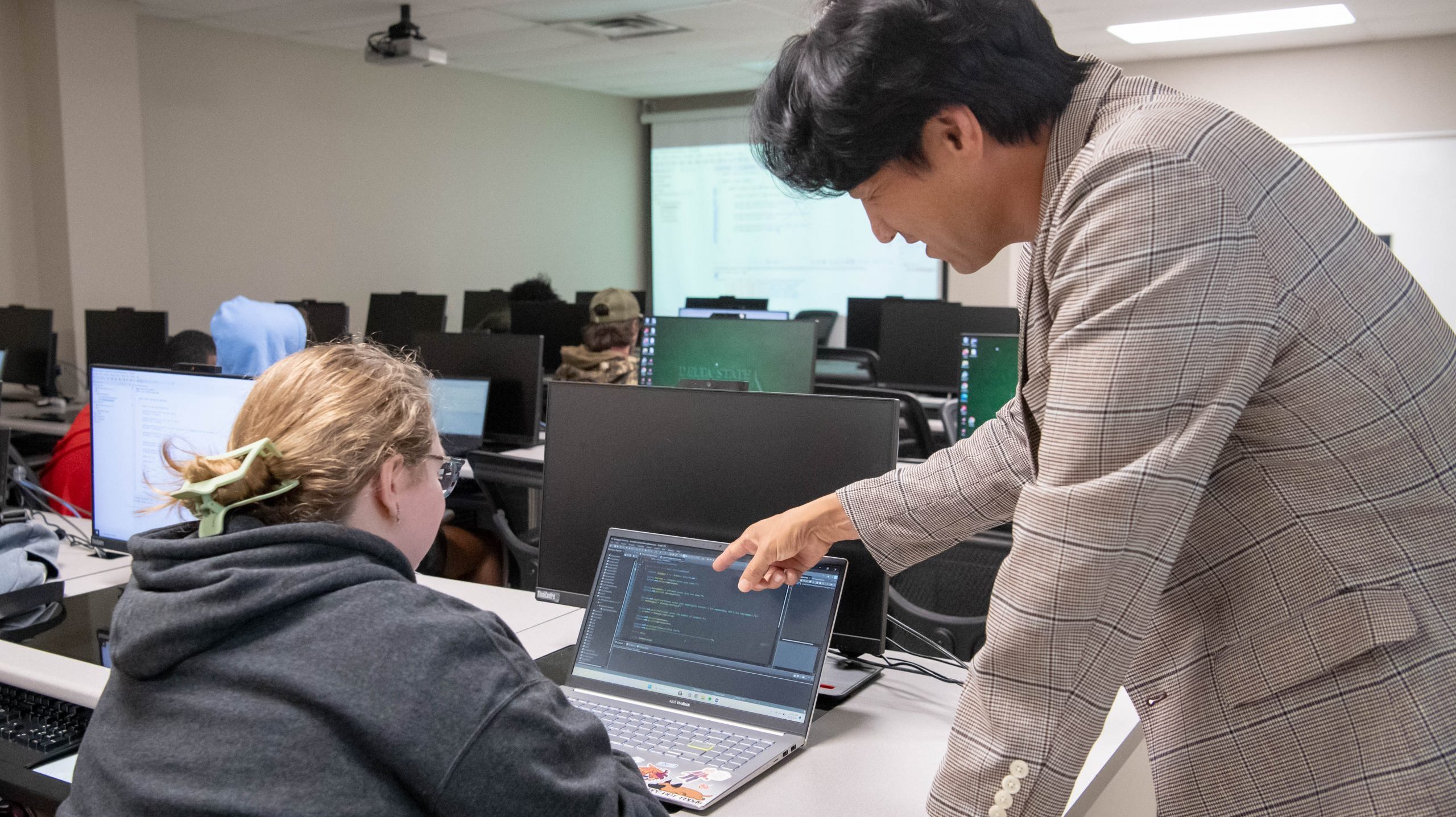 Professor in classroom pointing at code on students laptop screen.