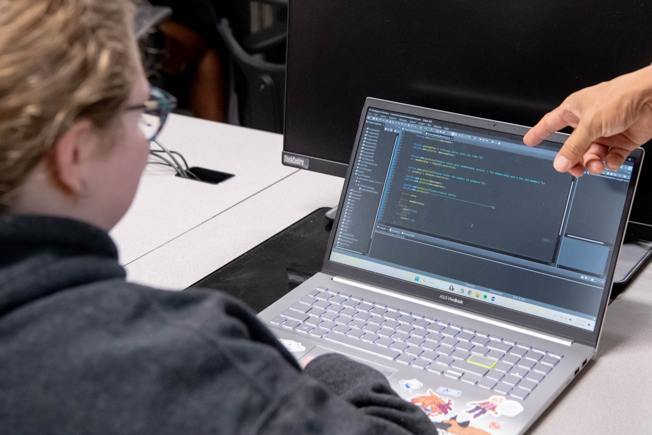 Student sitting in front of laptop with professors finger pointing at code on computer screen.