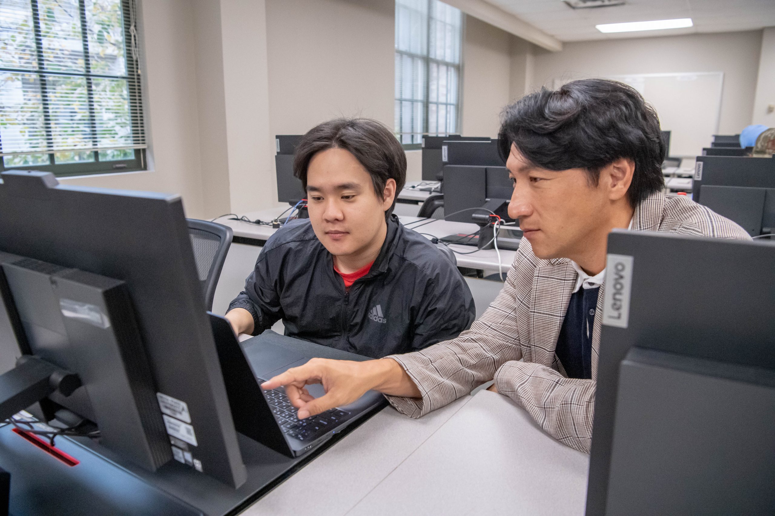 Professor sitting next to student in a computer lab, while pointing at students computer screen.