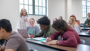 Students in a classroom listening and taking notes while a smiling instructor engages with the class.