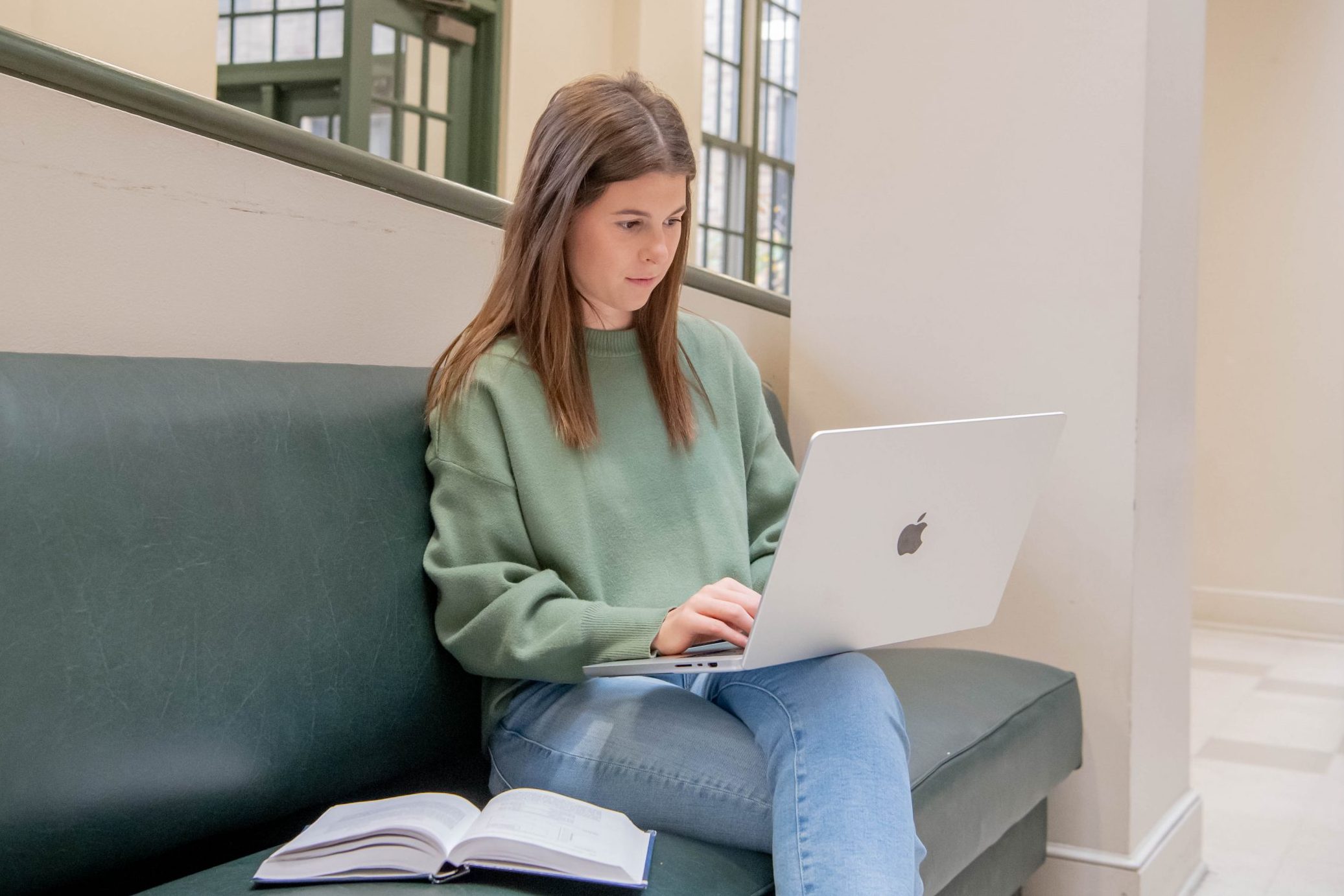 Student sitting on a bench with laptop in lap and open textbook on the bench in the College of Business lobby.