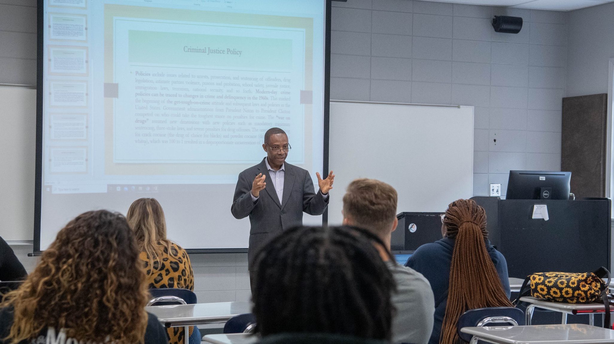 Professor giving lecture to class with PowerPoint projected behind him.