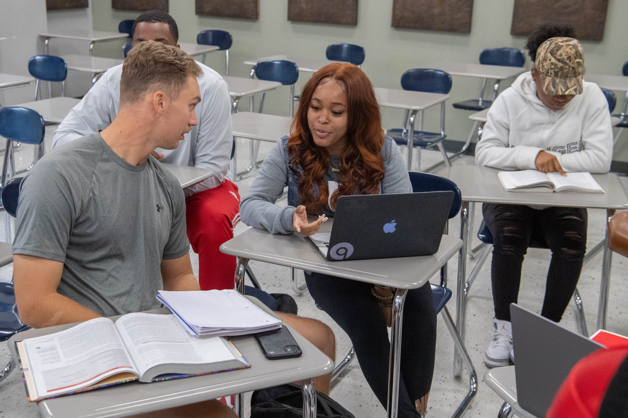 Four students sitting in a group in class, one with a laptop and one with a textbook and notebook.