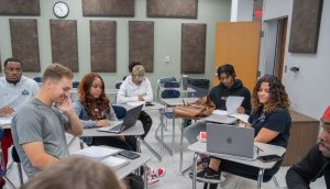 Seven students sitting in a group in class, with laptops and books.