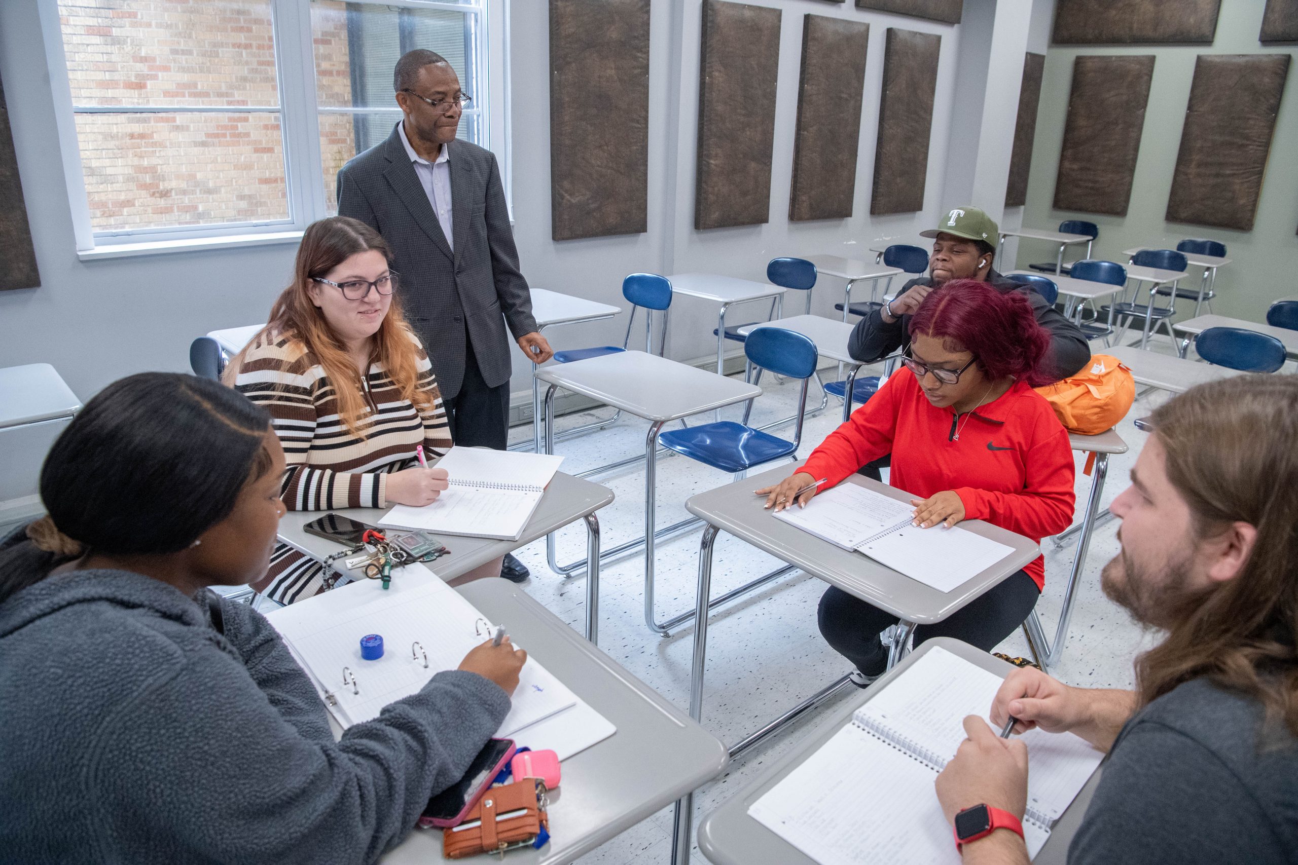 Five students with open notebooks sitting in a group in class, with a professor standing near them.