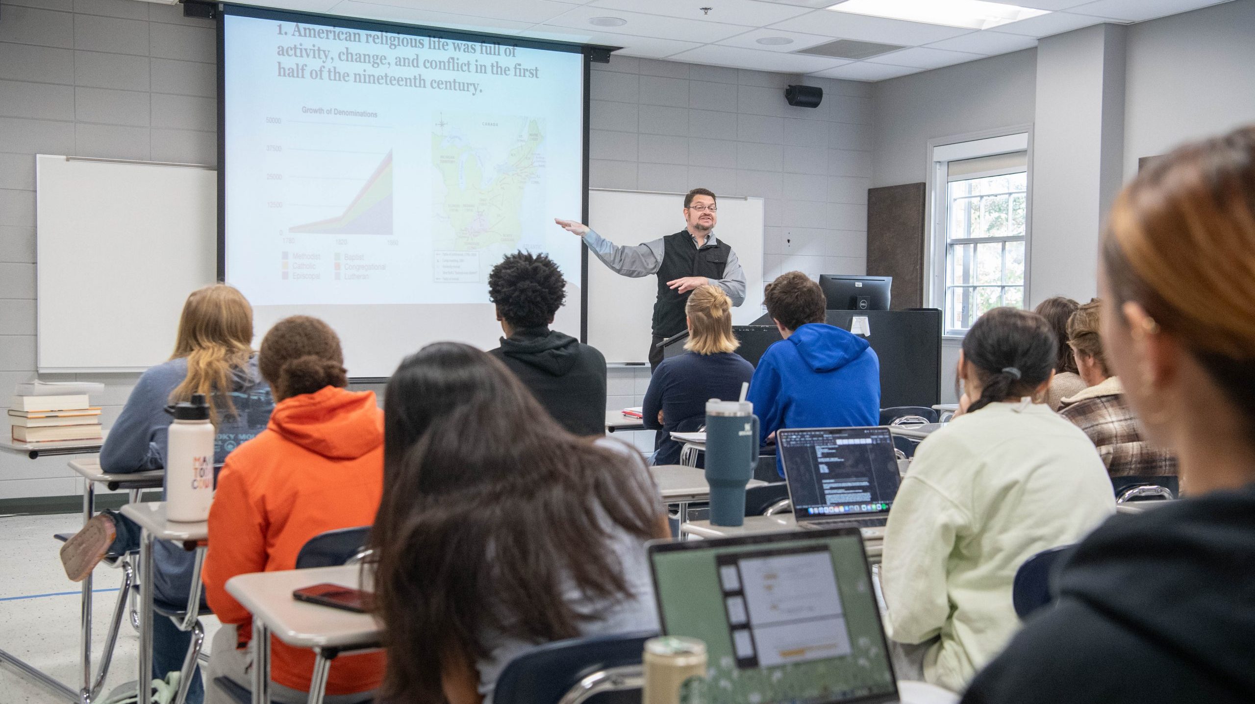 Professor giving lecture, with a PowerPoint next to him, in front of students in a classroom.