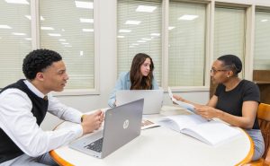 Three students sitting at a table with books and a laptop discussing a mock case.