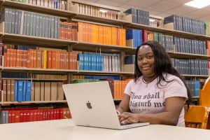 Student sitting at a table in the library on a laptop with bookshelves behind her.