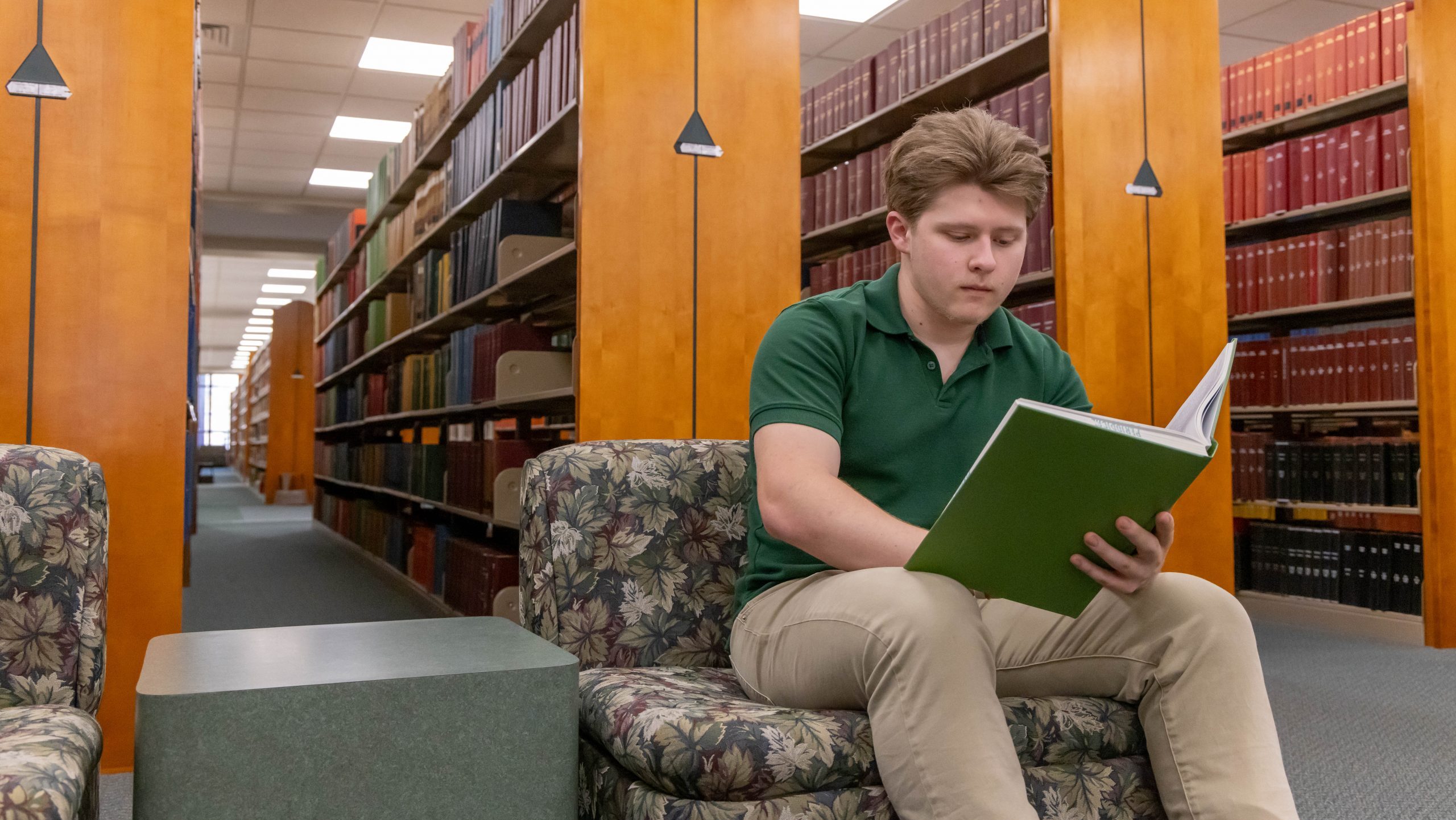 Student sitting near book shelves reading a book in the library.