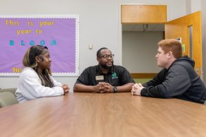 An Educational professional sitting at a table and having a conversation with two students, one on each side of him.