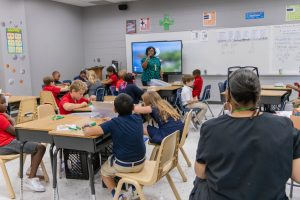 An elementary classroom filled with students sitting at desks, as the teacher teaches class and an administrator observes.
