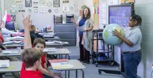 A teacher standing in front of the classroom with a student holding a globe and students in the classroom holding up their hands to answer a question.