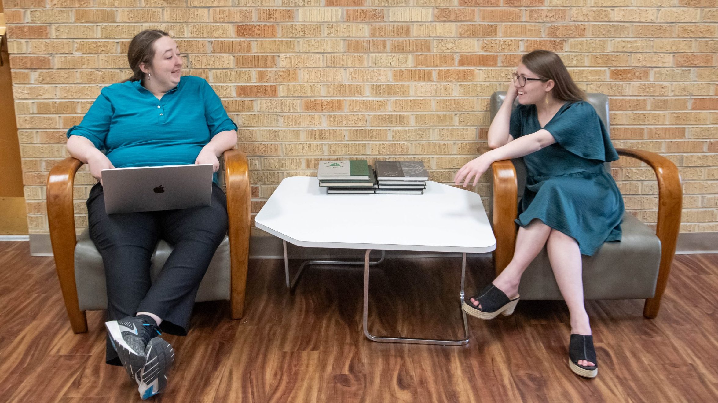 Two students having a conversation in a sitting area, one with a laptop.