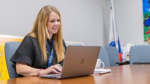 A woman sitting at a table in a conference room, smiling while looking at her computer.