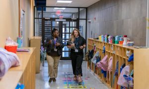 Two women talking while walking down an elementary school hallway that has cubbies filled with student belongings lining the walls.