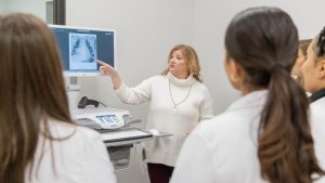Instructor points to a chest X-ray on a monitor while teaching a group of students in lab coats during a medical training session.