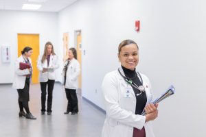 Smiling woman in a lab coat holding a clipboard in the foreground, with three other women in lab coats talking in the background.