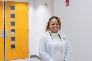 Smiling woman in a lab coat stands near bright yellow doors in hallway of academic medical setting.