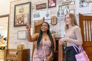 Two female students standing in a museum, one pointing toward an exhibit on the wall while the other student actively listens.