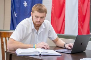 A man seated at a table, studying with one hand on an open book and the other on a laptop in front of him. There are flags in the background.