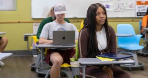 A high school girl sitting at a desk in a classroom with other students sitting at desks behind her.